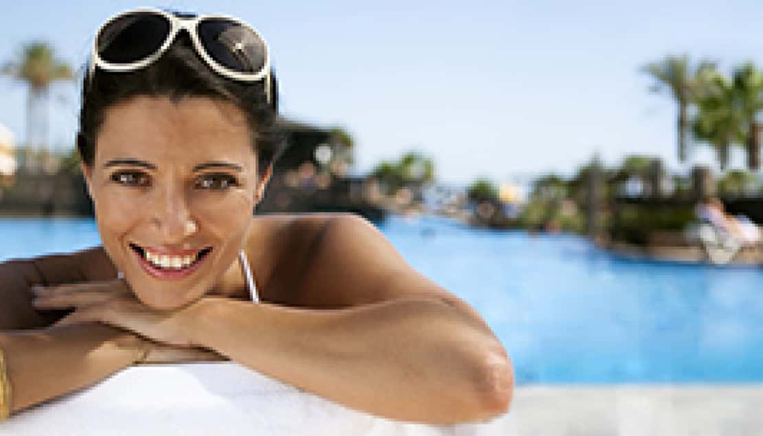 Smiling woman in sunglasses resting her arms on a pool edge, with palm trees and a resort behind.